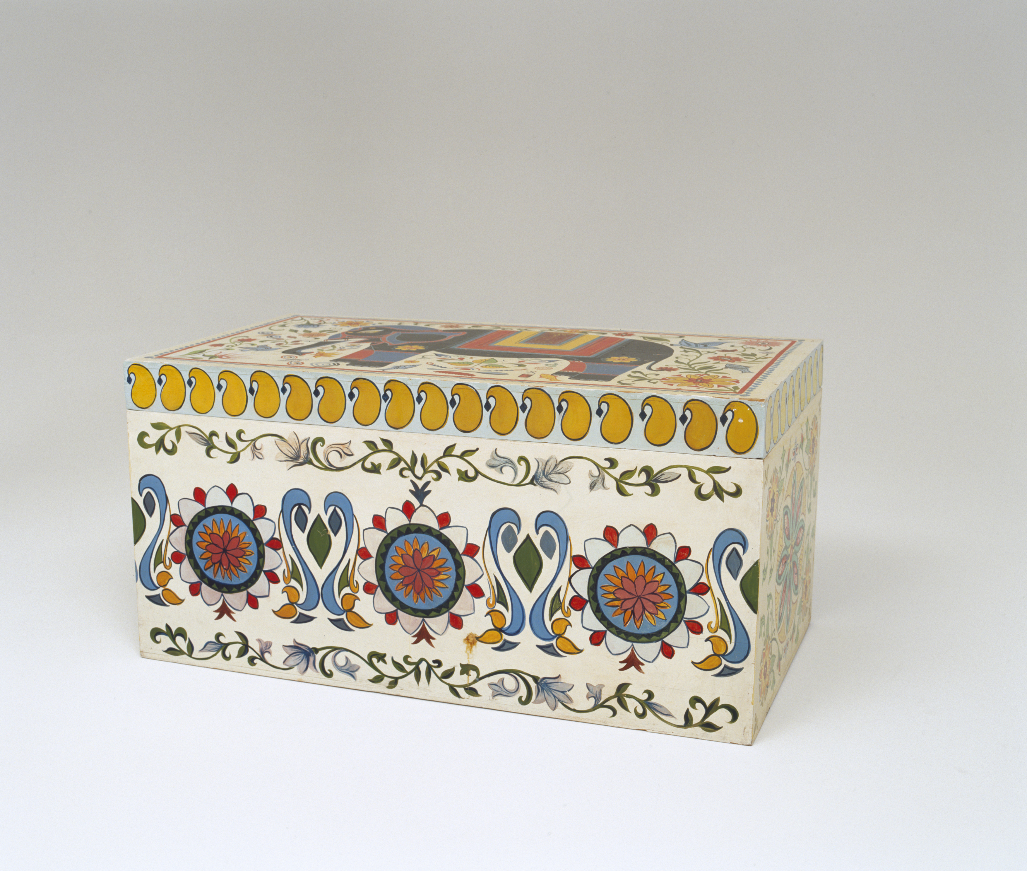 Wooden linen chest hand-painted to match the nakshi kantha. It is decorated on the top and all four sides. On the top is a black elephant with yellow red and blue trappings surrounded by flowers, leaves, fish and some articles of everyday usage. The border is a red line with a blue 'ribbon' pattern outside it. Each end of the box is decorated with a stylised lotus flower in the centre surrounded by more flowers, leaves and insects. On the front of the box is a whole row of stylised lotus flowers surrounded by swirling fronds of vegetation. There is a row of yellow boteh (in Bangladesh, kalka) around the top.